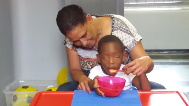 Helper behind child holding dominant hand with spoon, child's other hand on bowl, which  is placed on non-slip mat