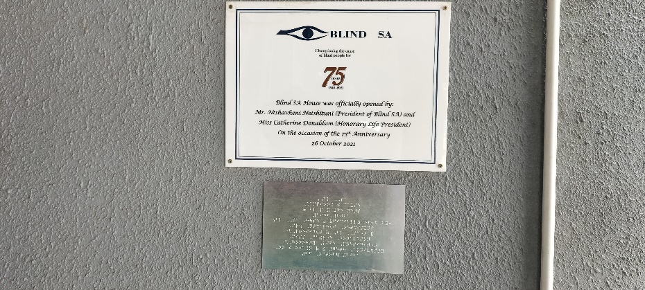 Photo of the plaque that is against the wall at the entrance of the office. It reads as follows: Blind SA House was officially opened by: Mr. Ntshavheni Netshituni (President of Blind SA) and Miss Catherine Donaldson (Honorary Life President) on the occasion of the 75th Anniversary 26 October 2022