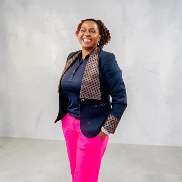 A smiling woman, the new CEO of Blind SA, stands confidently with one hand in her pocket. She wears a navy blazer with patterned trim, a navy shirt and bright pink trousers against a light grey background.